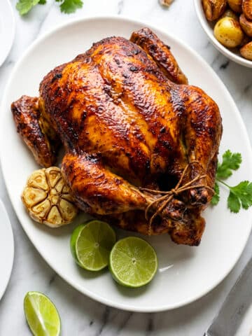 Pollo Rostizado (Rotisserie Chicken) served on a white platter with lime wedges, whole roasted garlic and cilantro garnish.