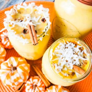 Pumpkin spice coquito in mason jars topped with chopped walnuts and coconut flakes. Small foam pumpkins surrounding the jars.