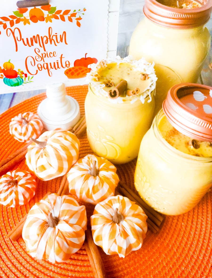 Pumpkin spice coquito in mason jars topped with chopped walnuts and coconut flakes. Small foam pumpkins surrounding the jars.