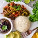 Ropa Vieja served on a white plate with a side of white rice, black stewed beans and avocado slices.
