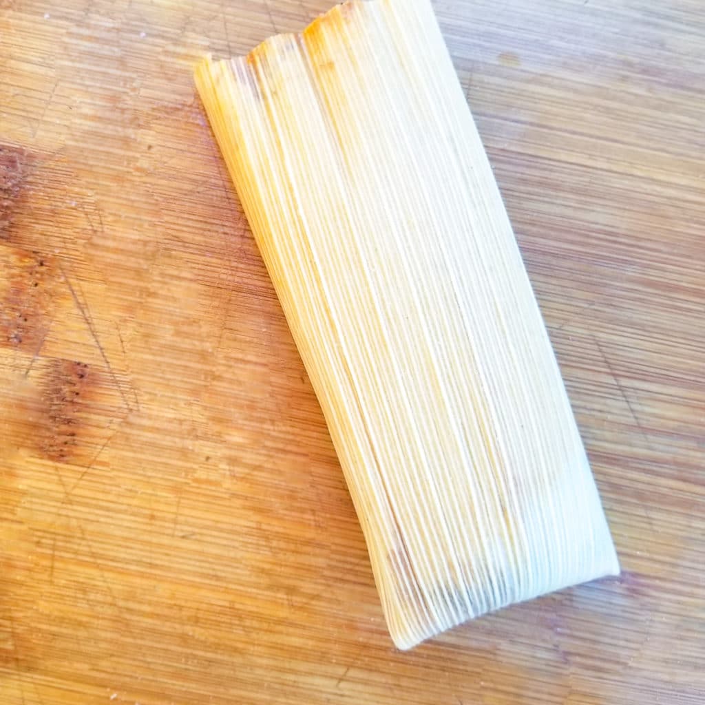 Folding tamale on a wooden cutting board.