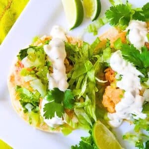 Tostadas de Camaron topped with shredded lettuce, jalapeno tartar sauce, tomatillo pico de gallo and cilantro, served on a white platter with lime wedges.