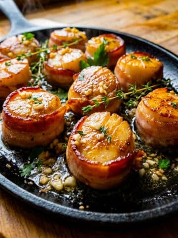 Vieras de Mar, bacon wrapped sea scallops cooked in an oval cast iron skillet, topped with a butter garlic sauce and garnished lightly with parsley and thyme.